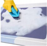 Osoba w żółtej rękawicy czyści powierzchnię szczotką, używając detergentu z obfitą pianą.