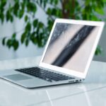 Laptop na biurku z rośliną w tle. Ekran rozjaśniony, nowoczesna technologia w biurowym otoczeniu.