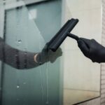 Osoba w czarnych rękawiczkach czyści szybę ściągaczką, usuwając krople wody.