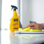 Butelka płynu do czyszczenia ekranów LCD obok osoby wycierającej powierzchnię szmatką w jasnym pomieszczeniu.