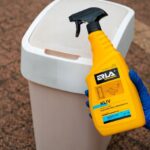 Spray do czyszczenia plastiku trzymany w ręku z rękawiczką, obok plastikowego pojemnika na zewnątrz.