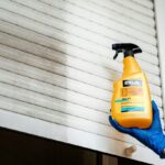 Środek czyszczący do żaluzji ERLA, trzymany w ręce w niebieskiej rękawiczce, na tle zabrudzonej żaluzji.