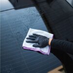 Osoba w rękawiczce przeciera mokrą szybę samochodu ściereczką, zapewniając czystość i połysk.