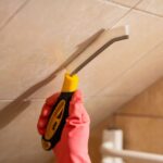 Ręka w rękawiczce trzyma szczotkę do czyszczenia fug na płytkach łazienkowych.