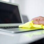 Ręka czyści ekran laptopa żółtą ściereczką, dbając o czystość urządzenia elektronicznego w biurze.