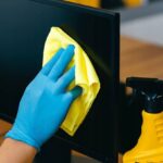 Ręka w niebieskiej rękawiczce czyszcząca ekran monitora żółtą ściereczką z mikrofibry obok butelki ze środkiem czyszczącym.