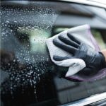 Czyszczenie mokrej szyby samochodowej ręcznikiem z mikrofibry przez osobę w czarnej rękawiczce.