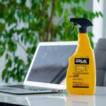 Spray do czyszczenia ekranów obok laptopa na biurku, w tle zielone liście rośliny.