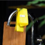 Żółta ściereczka z mikrofibry na chromowanym kranie w kuchni z deską do krojenia w tle, ERLA Home.