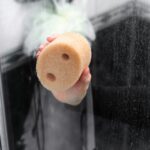 Ręka trzymająca gąbkę do czyszczenia w łazience z zaparowaną szybą prysznicową w tle.