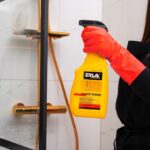 Osoba czyści kabinę prysznicową płynem ERLA Saniz Shine w żółtej butelce, mając na sobie czerwone rękawice.