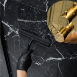 Czarna rękawica na dłoni trzyma plastikową ściągaczkę do wody na marmurowych, czarnych kafelkach w łazience.