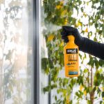 Osoba w rękawiczce trzyma żółty spray do czyszczenia szyb na tle okna i zielonych liści.