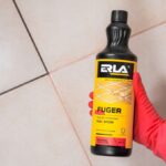 Płyn do czyszczenia fug ERLA w czarnej butelce, trzymanej przez rękę w czerwonej rękawicy na tle płytek.