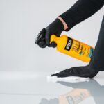 Osoba w czarnych rękawiczkach używa żółtego sprayu czyszczącego do polerowania powierzchni ściereczką.