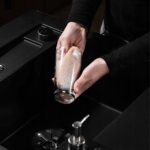 Osoba myjąca szklankę w kuchennym zlewie, używając gąbki i środków do czyszczenia naczyń.