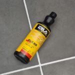 Płyn do czyszczenia fug na tle szarej podłogi — ERLA Fuger, żółto-czarna butelka.