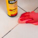 Ręka w rękawiczce czyści fugę kafelki płynem do czyszczenia Erlax Fuger i czerwoną ściereczką.