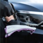 Osoba spryskuje ręcznik mikrofibrowy środkiem czyszczącym przed polerowaniem auta, zbliżenie na reflektor.