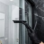 Czyszczenie szklanej kabiny prysznicowej gumową ściągaczką, czarne rękawiczki, marmurowe tło łazienkowe.